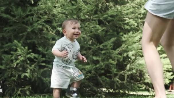 Ein kleiner Junge in T-Shirt und kurzer Hose tanzt auf dem Rasen im Garten neben seiner jungen Mutter. Zeitlupe. - Filmmaterial, Video