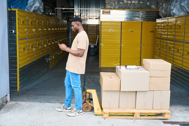Hombre afroamericano hablando por teléfono, apoyado en el carro de carga, en el carro hay cajas de cartón y un dispensador de cinta - Foto, Imagen