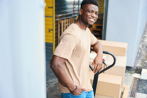 African American guy leaning against a cargo cart, there are cardboard boxes and a tape dispenser on the cart - Photo, Image