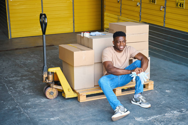 African American man sat down to rest on cargo cart, on the cart are cardboard boxes and a tape dispenser - Photo, Image
