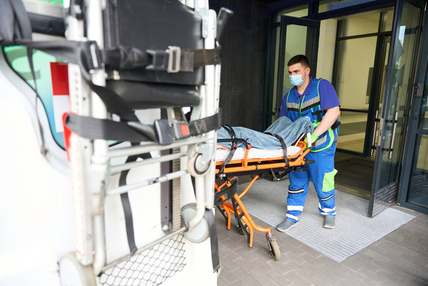 Záchranář v ochranné masce vyloží nosítka s pacientem ze sanitky, pacienta odvezli na pohotovost. - Fotografie, Obrázek