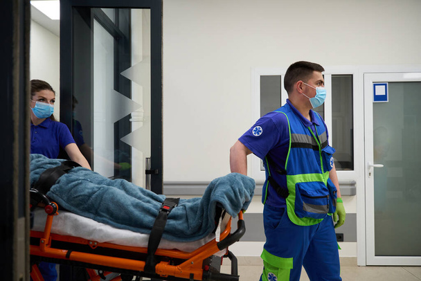 Sanitariusze przynoszą wózek z pacjentem na korytarz szpitalny, personel medyczny pracuje w maskach ochronnych. - Zdjęcie, obraz