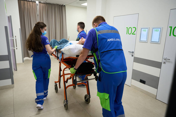 Команда врачей несет каталку с пациентом вдоль больничного коридора, пациент был поставлен на капельницу с обезболивающими - Фото, изображение