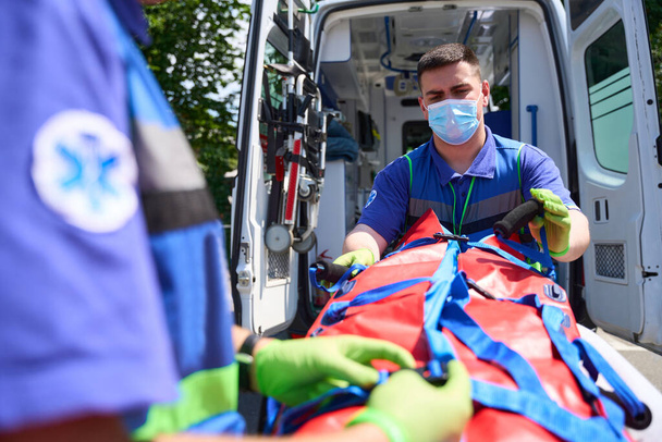 Чоловічі медики фіксують жертву в аварії на спеціальному носику, парамедики в захисних масках і рукавичках - Фото, зображення