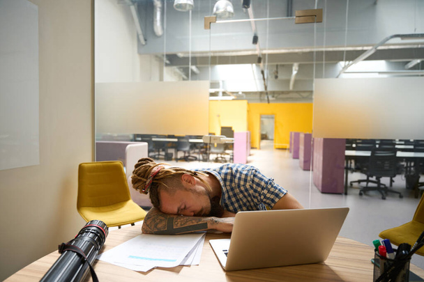 Freelancer in een geruit shirt zit aan zijn bureau ingedommeld tijdens het werk als gevolg van slechte gezondheid - Foto, afbeelding