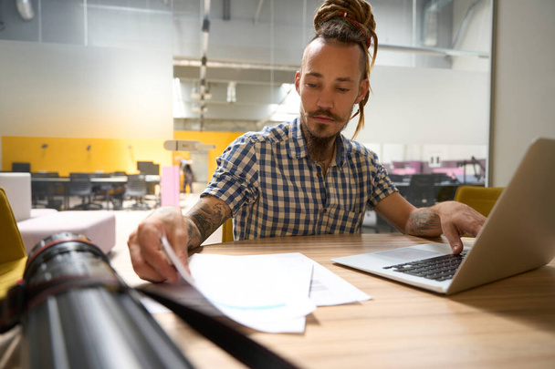 Ekose gömlekli bir adam çalışma kağıtları, bir dizüstü bilgisayar ve masanın üzerinde çizimler olan bir tüple çalışır. - Fotoğraf, Görsel