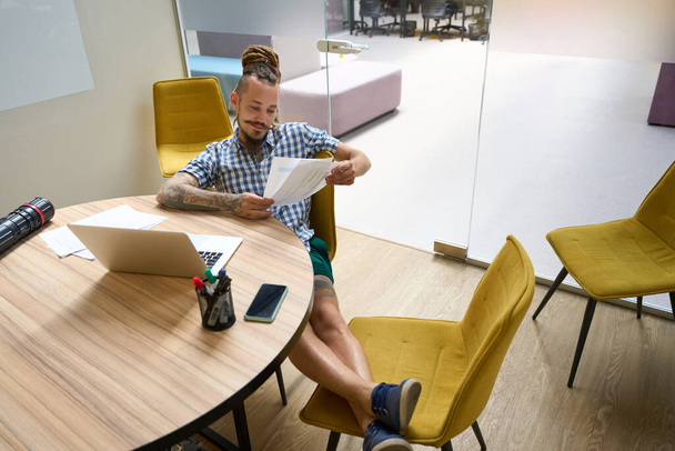Ekose gömlekli adam çalışma kağıtları, dizüstü bilgisayar ve masanın üzerinde çizimler olan bir tüple masada oturuyor. - Fotoğraf, Görsel