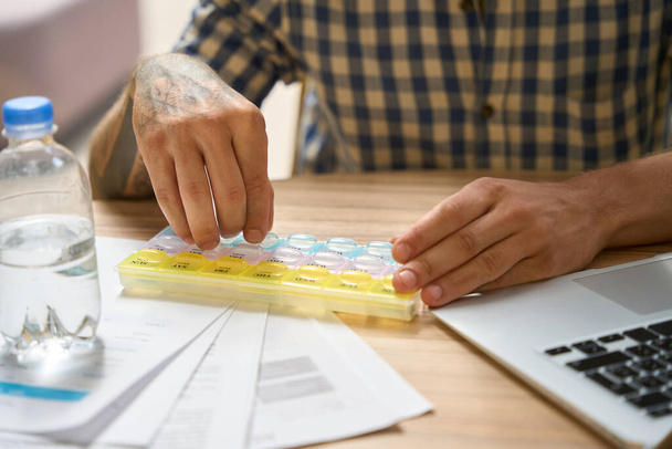 Мужчина в клетчатой рубашке выкладывает таблетки в клетках специального органайзера для таблеток - Фото, изображение