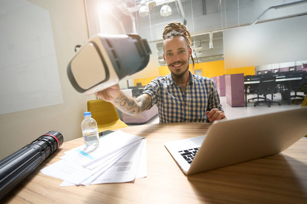 Fiatal srác tartja virtuális valóság headset a kezében, ő ül az asztalánál egy coworking térben - Fotó, kép