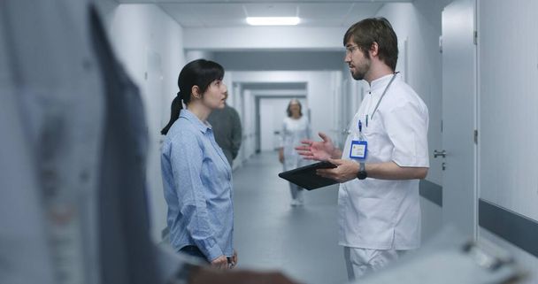 Мужчина-врач стоит в коридоре клиники с пациенткой, использует цифровые таблетки. Профессиональный медик беседует со взрослой женщиной, обсуждает результаты медицинских тестов. Медицинский персонал и пациенты в коридоре больницы - Фото, изображение