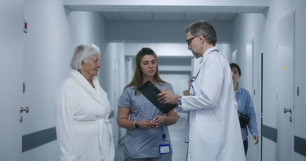 Зрілий лікар стоїть в клінічному коридорі з медсестрою і пацієнтом. Професійні медичні розмови з колегою і літньою жінкою, використовують цифровий планшет. Медичний персонал та пацієнти в лікарняному коридорі. - Фото, зображення