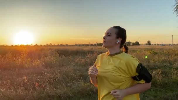 電話アームバンドを使用してスポーツウェアの若い女性は,素晴らしい日没の間に美しい田舎の風景の中でワイヤレスイヤホンを介して音楽を聞いている間,彼女の夜のジョギングを楽しんでいます - 映像、動画