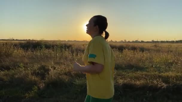 Zijaanzicht jonge vrouw in levendige sportkleding joggen langs serene landweg, omgeven door het pittoreske landelijke landschap, badend in het gouden avondzonlicht. Trainen routine, actieve levensstijl - Video
