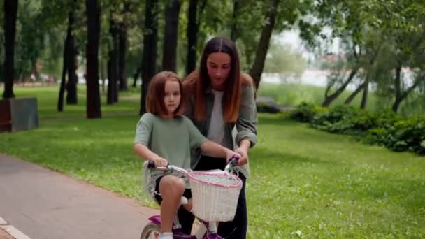 retrato familia joven madre enseñando hija a montar en bicicleta en ciudad parque concepto feliz infancia y cuidado - Imágenes, Vídeo