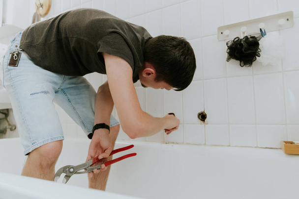 Un giovane caucasico riconoscibile ragazzo con una bruna usa il dito per pulire un buco da un rubinetto in una parete piastrellata bianca in bagno a casa, vista laterale da vicino. - Foto, immagini