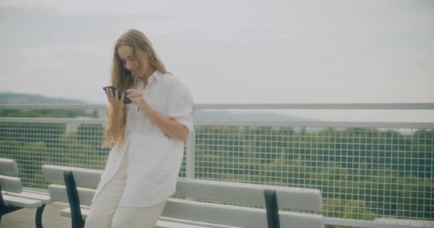 スマートフォンを使用して笑顔の若い女性のスローモーションショットは,レイリングの近くのベンチに傾いています - 映像、動画