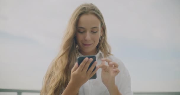 Fotografía en cámara lenta de una atractiva joven con el pelo rubio disfrutando mientras usa el teléfono inteligente contra el cielo - Metraje, vídeo