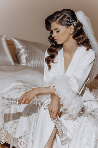 Piękna młoda panna młoda z długimi kręconymi włosami w modnej białej szacie siedzi na łóżku w hotelu i trzyma okulary w rękach. Młoda piękna dziewczyna rano w białej sukience. - Zdjęcie, obraz