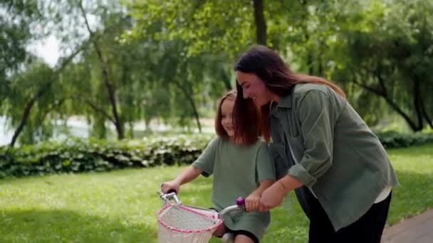 família jovem mãe ensinando filha a andar de bicicleta no parque da cidade conceito de infância feliz e cuidado - Filmagem, Vídeo