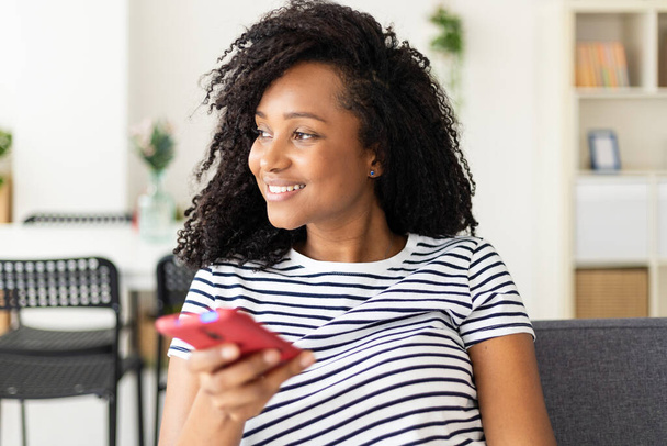Happy young adult african american woman holding phone sitting on sofa at home - Фото, зображення