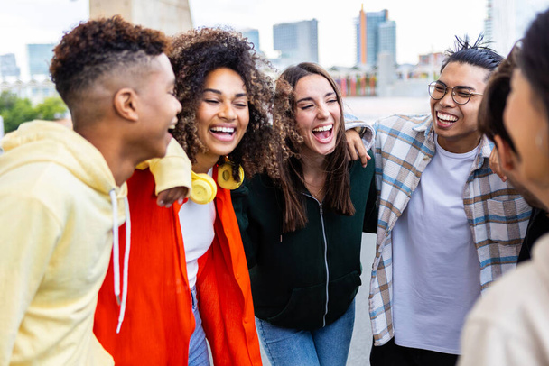 Young group of happy students hugging each other laughing together outside. College diverse friends laughing enjoying lifestyle moment standing in city street. Youth community and friendship concept. - Photo, Image