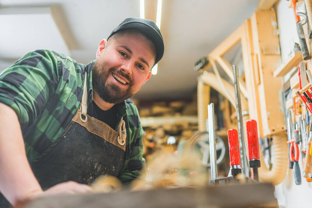 Πορτρέτο του χαμογελαστού καυκάσιου ξυλουργού στο ξυλουργικό του τραπέζι. Υψηλής ποιότητας φωτογραφία - Φωτογραφία, εικόνα