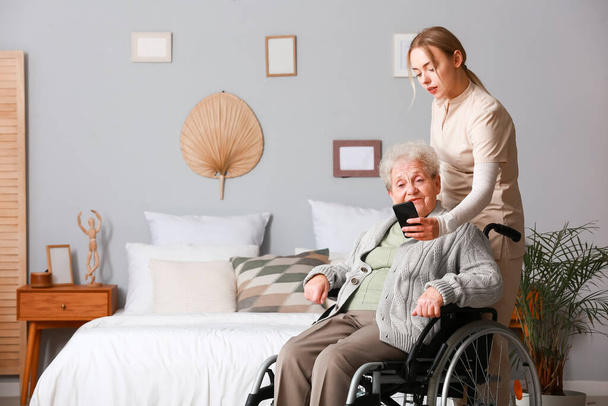 Νεαρός φροντιστής με ηλικιωμένη γυναίκα σε αναπηρική καρέκλα που χρησιμοποιεί κινητό τηλέφωνο στο σπίτι - Φωτογραφία, εικόνα