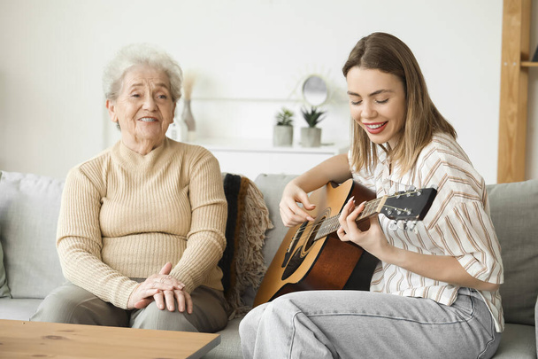シニアの女性と孫娘が自宅でギターを弾く - 写真・画像
