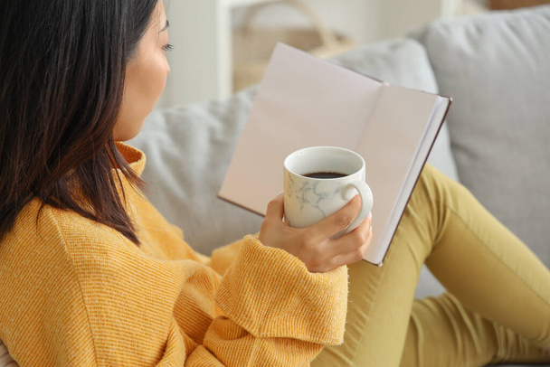 Όμορφη νεαρή Ασιάτισσα γυναίκα με φλιτζάνι τσάι κάθεται στον καναπέ στο σαλόνι και το βιβλίο ανάγνωσης - Φωτογραφία, εικόνα
