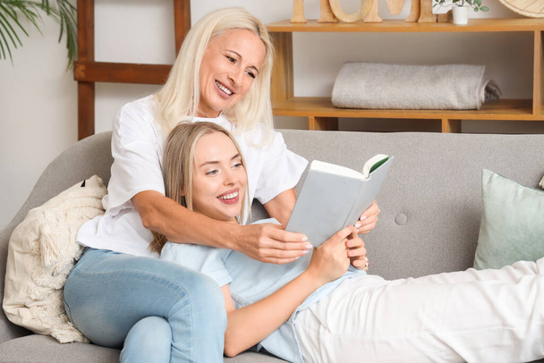 Young beautiful woman with her mother reading book together in living room - Foto, Imagem