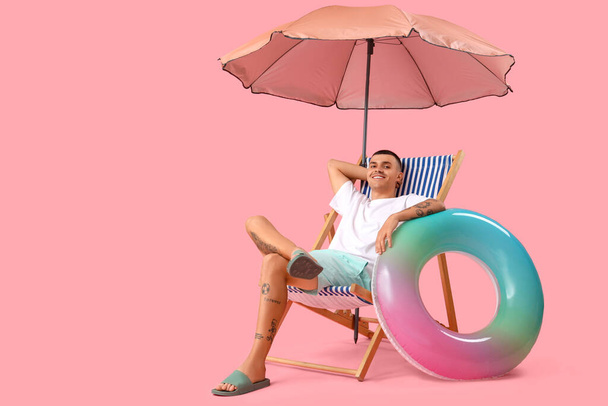 Szczęśliwy młody człowiek z nadmuchiwanym pierścieniem i parasolem siedzący na leżaku na różowym tle - Zdjęcie, obraz