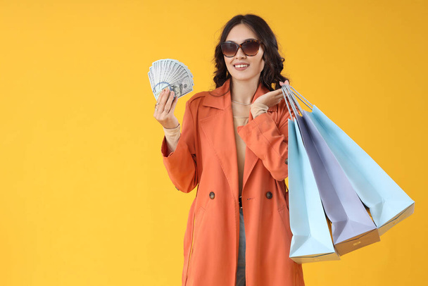 Young Asian woman in fall clothes with money and shopping bags on yellow background - Photo, Image