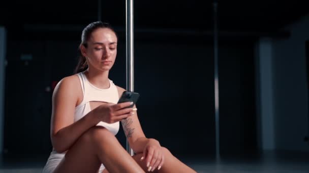 retrato de uma jovem bela menina sexy depois de uma aula de dança em um poste segurando telefone em suas mãos mensagens de texto com alguém - Filmagem, Vídeo