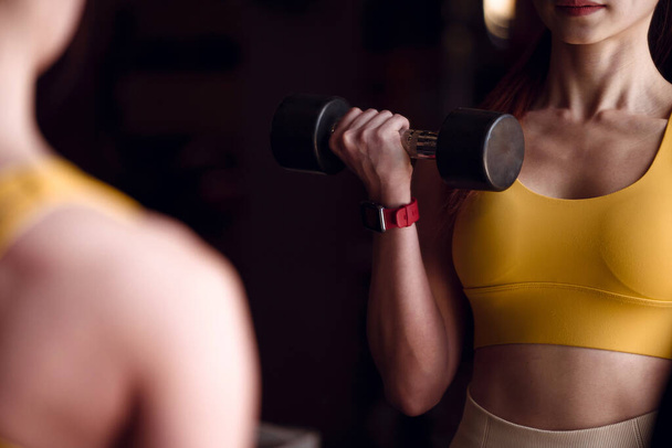 Young female doing exercise with dumbbell in front of a mirror in the gym. - Photo, Image