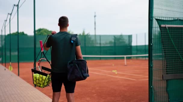 entrenador joven jugador profesional de pie en la cancha de tenis al aire libre antes de comenzar el entrenamiento del juego con raqueta y balones de tenis canasta vista posterior - Imágenes, Vídeo