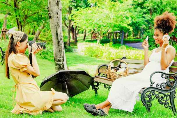 Asijské přítelkyně fotograf sedí a fotografování focení, portrét zdravé těhotné africké Američanky sedí na dřevěné lavičce v parku ukazuje rukavice dítě usmívá se na kameru šťastný. - Fotografie, Obrázek
