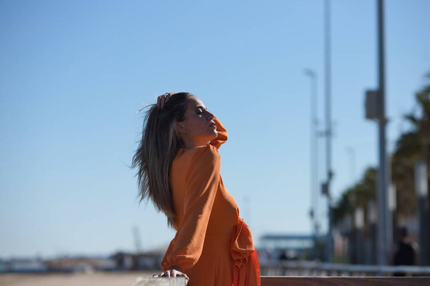 Retrato de perfil de una guapa joven rubia vestida de naranja apoyada sobre una barandilla de madera en la playa. Cielo azul en el fondo en el horizonte. Concepto de moda y belleza - Foto, imagen