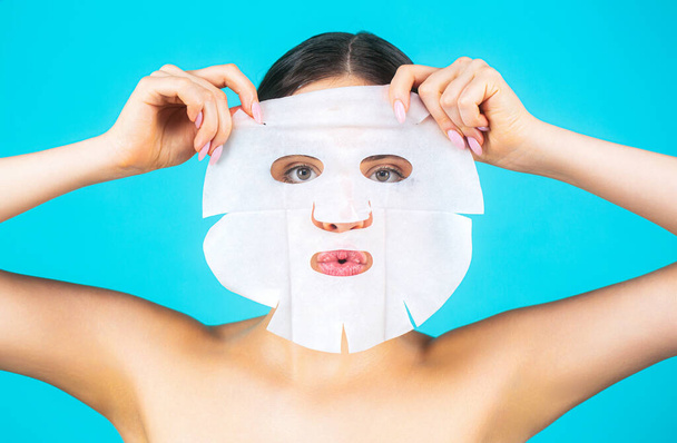 Beautiful woman with mask. Girl taking care of skin complexion. Skin care and beauty concept. Moisturizing mask. Woman applying sheet mask on her face, on blue background. - Photo, Image