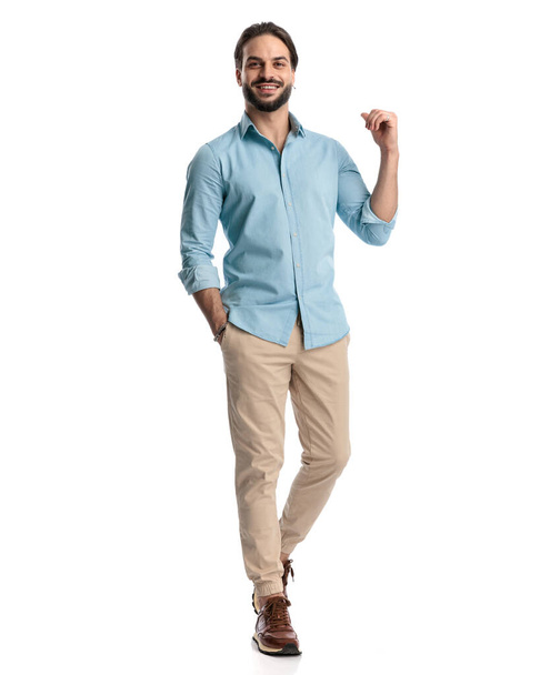 sexy mladý muž s vousy chůze s rukou v kapse a pěst nahoru a usmívat se před bílým pozadím - Fotografie, Obrázek