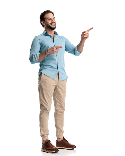 attraktiver junger Mann mit Bart lächelt, während er die Finger wegzeigt und vor weißem Hintergrund posiert  - Foto, Bild