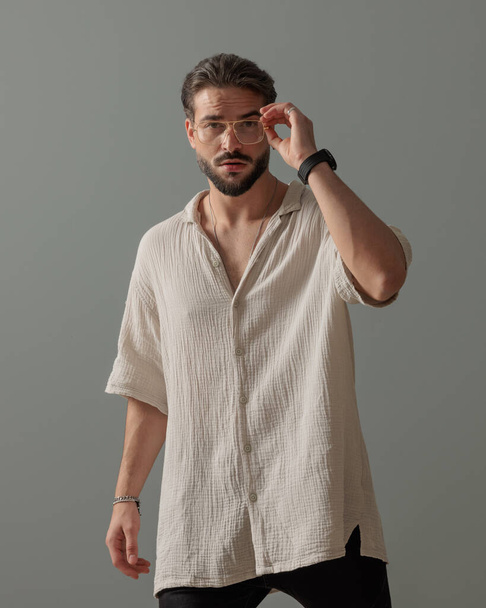 Cooler Modemann blickt nach vorn und arrangiert Brille vor grauem Hintergrund  - Foto, Bild