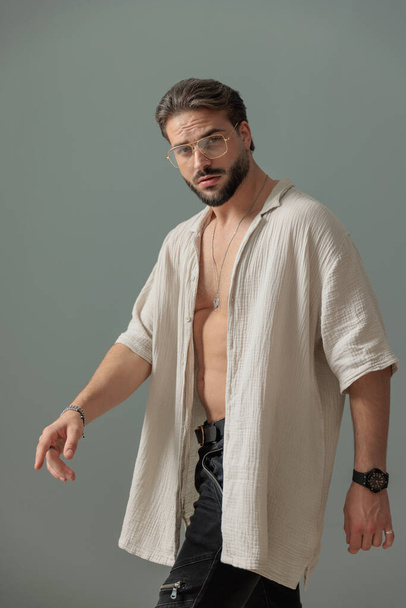 vonzó fiatal férfi szemüveges séta nyitott ing és magabiztos előtt szürke háttér - Fotó, kép