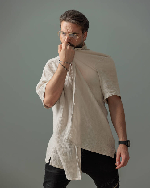 moda chico con gafas mirando hacia otro lado mientras cubre la cara con camisa en frente de fondo gris  - Foto, Imagen