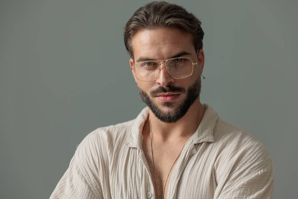 portré szexi szakállas fickó szemüveges előretekintő és pózol előtt szürke háttér - Fotó, kép