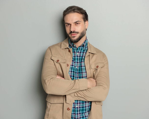 handsome bearded casual man in jacket crossing arms and looking forward in front of grey background - Photo, Image