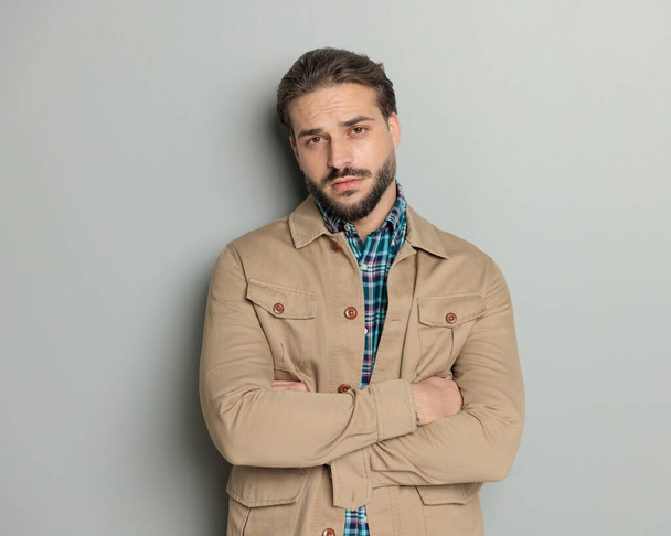 портрет сексуального бородатого мужчины в бежевой куртке, скрещивающего руки и позирующего, глядя вперед на сером фоне  - Фото, изображение