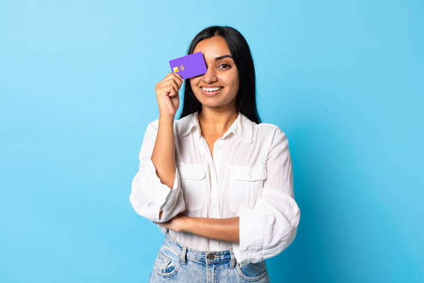 Grande oferta bancária. Jovem alegre indiana senhora segurando cartão de crédito bancário fechando os olhos, sorrindo para a câmera ao anunciar o serviço financeiro, de pé sobre fundo azul estúdio - Foto, Imagem