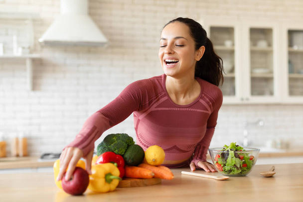 Glückliche junge, sportliche Frau, die in der Küche gesunden Gemüsesalat zubereitet, genießt es, zu Hause zu kochen, frisches Obst und Gemüse zu sich zu nehmen und das Abendessen zuzubereiten. Ernährung bei Gewichtsverlust - Foto, Bild