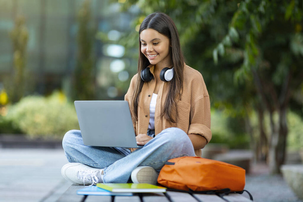 Online koulutus, opiskelijoiden elämäntapa käsite. Iloinen viehättävä nuori nainen opiskelija tekee kotitehtäviä puistossa, istuu penkillä, kannettavan tietokoneen, langattomat kuulokkeet, kopioi tilaa - Valokuva, kuva