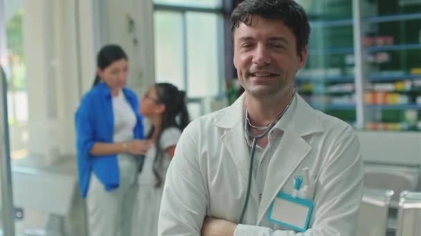 Porträt eines fröhlichen Kinderarztes mit Stethoskop, der mit Frau und krankem Mädchen im Hintergrund in der Klinik steht und in die Kamera blickt - Filmmaterial, Video